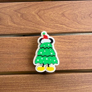 May include: A flat, die-cut acrylic charm shaped like a green Christmas tree with white polka dots and small white mouse-ear silhouettes. The tree wears a red and white Santa hat at the top and has black mouse ears, thin black legs, and yellow shoes.