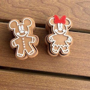 May include: Two brown gingerbread-style cookie cutters shaped like cartoon mice. The left cutter features a mouse with white icing details. The right cutter features a mouse with a red bow and white icing details, including a dress pattern.