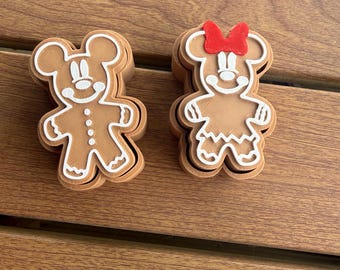 May include: Two brown gingerbread-style cookie cutters shaped like cartoon mice. The left cutter features a mouse with white icing details. The right cutter features a mouse with a red bow and white icing details, including a dress pattern.