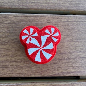 May include: A red and white decorative charm shaped like a stylized head with three circular sections. The surface features a peppermint candy pattern with radiating stripes in alternating red and white colors across the entire front face.