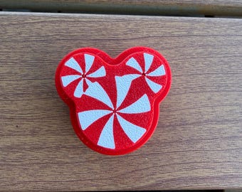 May include: A red and white decorative charm shaped like a stylized head with three circular sections. The surface features a peppermint candy pattern with radiating stripes in alternating red and white colors across the entire front face.