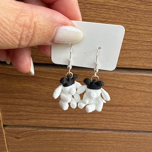 May include: A pair of drop earrings featuring small, embroidered figures with white bodies and black heads. The figures are attached to silver-toned metal fishhook wires and are displayed on a rectangular white card for presentation.