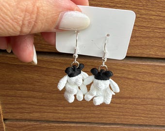 May include: A pair of dangle earrings featuring small, embroidered figures with white bodies and black heads. The figures are attached to silver-toned metal fishhook wires and are displayed on a rectangular white card for presentation.