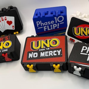 Peut inclure: Six boîtiers de jeux de cartes rectangulaires en plastique avec divers designs, notamment UNO, Phase 10 et Liar's UNO. Les boîtiers sont noirs, blancs et bleus avec des logos colorés et du texte comme Show 'Em No Mercy, Trust No One et Phase 10 Flip.