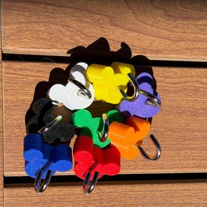 May include: A collection of seven colorful hooks, each featuring a mouse-ear silhouette design. The hooks are available in white, yellow, purple, orange, red, blue, green, and black, with each piece including a curved silver metal hook attachment.