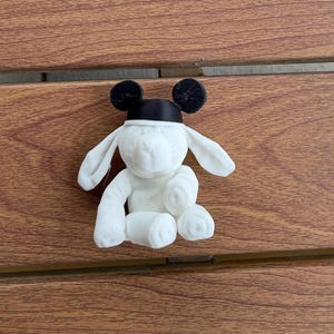 May include: A white plush toy shaped like a dog with long floppy ears and a black headband featuring two round black ears on top. The toy has a soft, textured surface and is positioned in a seated posture with its front paws resting forward.