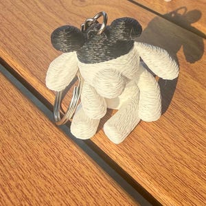 May include: A 3D printed keychain featuring a stylized animal figure with a black head and ears, and a white body. The figure has a textured, layered surface appearance and is attached to a circular silver metal split ring for keys.