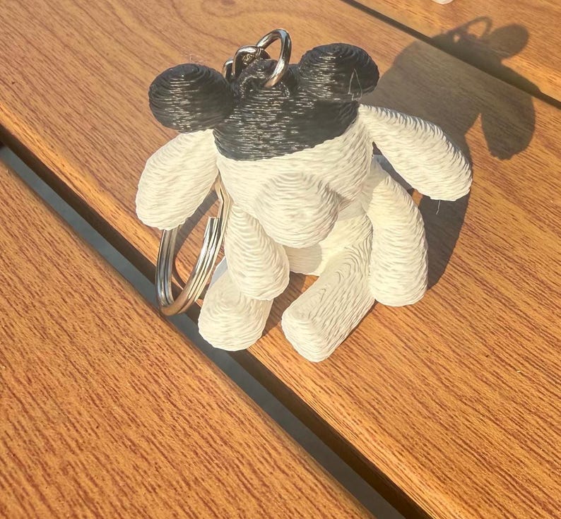 May include: A 3D printed keychain featuring a stylized animal figure with a black head and ears, and a white body. The figure has a textured, layered surface appearance and is attached to a circular silver metal split ring for keys.