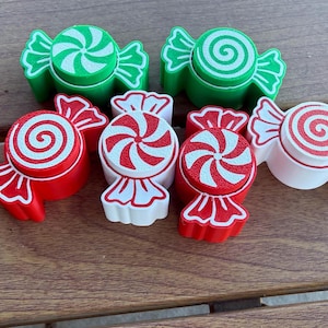 May include: A collection of six candy-shaped ornaments in red, green, and white. Each piece features a central circular swirl pattern and flared ends. The set includes two green, two red, and two white items, all with contrasting colored swirl designs.