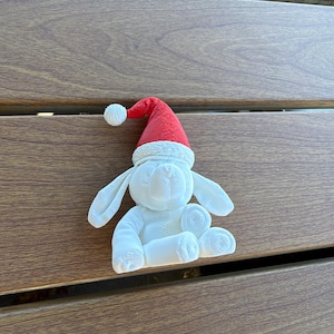 May include: A white rabbit figurine with long ears and a textured surface, wearing a bright red Santa hat with a white pom-pom and a white brim. The rabbit is in a seated position with its front paws resting forward and its ears hanging down.