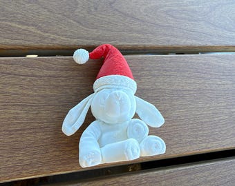May include: A white rabbit figurine with long ears and a textured surface, wearing a bright red Santa hat with a white pom-pom and a white brim. The rabbit is in a seated position with its front paws resting forward and its ears hanging down.