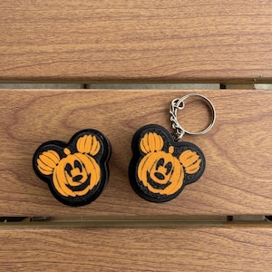 May include: Two black and orange pumpkin-shaped charms featuring a stylized mouse head silhouette. One charm is plain, while the other is attached to a silver-toned metal split ring keychain. The design includes carved facial features.