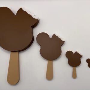 May include: A set of four brown Mickey Mouse-shaped ice cream bars on tan sticks, with a white bite taken out of the top of two. The sizes vary, with the largest measuring approximately 12 inches tall.
