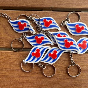 May include: A collection of keychains featuring a white, wave-shaped design with three blue stripes and a red Mickey Mouse head silhouette. Each keychain includes a silver-toned metal split ring attached by a short chain, showcasing a nautical theme.