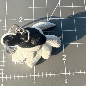 May include: A white and black 3D printed keychain shaped like a stylized creature with a textured surface. The keychain features a metal ring attached to the top. It is positioned on a gray cutting mat with visible white inch markings.