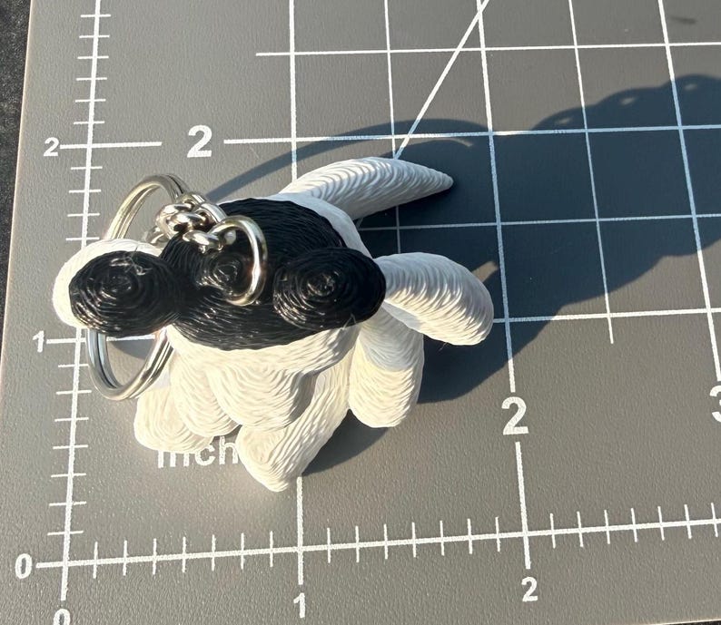 May include: A white and black 3D printed keychain shaped like a stylized creature with a textured surface. The keychain features a metal ring attached to the top. It is positioned on a gray cutting mat with visible white inch markings.