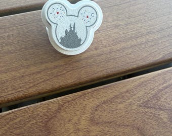 May include: A white circular phone grip featuring a gray Mickey Mouse head silhouette. Inside the ears are gray firework patterns with small red accents, and the center displays a gray castle silhouette against a textured white background.