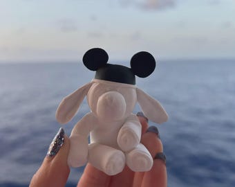 May include: A small white figurine of a dog with long floppy ears, wearing a black cap with two rounded black ears on top. The dog is in a seated position with its front paws resting on its lap, featuring a smooth matte finish throughout.