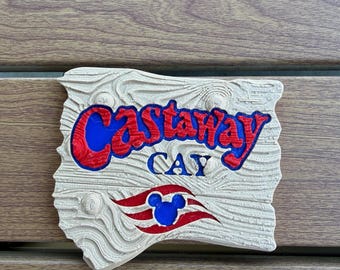 May include: A rectangular sign with a textured, light-colored wood grain finish. The text Castaway CAY is embossed in red and blue. Below the text is a red and white wavy graphic featuring a blue Mickey Mouse icon in the center.