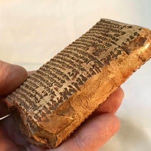 Puede incluir: Un artefacto rectangular de aspecto antiguo, con una base marrón claro y marcas marrones oscuras. El objeto, sostenido en una mano, muestra una superficie texturizada y un aspecto envejecido. Las marcas parecen ser una inscripción.