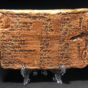 May include: A rectangular, weathered clay tablet with cuneiform writing. The tablet is a warm brown color and is supported by a clear plastic stand. The surface is textured with visible indentations from the writing process.