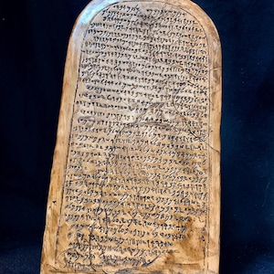 Puede incluir: Una tableta rectangular de color marrón claro con bordes superiores redondeados. La superficie está cubierta de texto oscuro escrito a mano. La tableta tiene una apariencia agrietada, lo que sugiere antigüedad. El texto parece ser una escritura antigua.