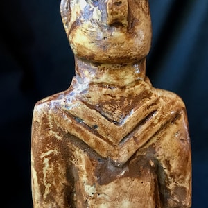 May include: A ceramic sculpture of a figure with a weathered appearance. The statue has a light brown base colour with darker brown accents and a textured surface. The figure has a stylized face and a geometric design on the chest.