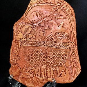 May include: A reddish-brown stone carving with detailed relief work. The rectangular piece shows figures, animals, and patterns, possibly representing a historical or mythological narrative. The carving is displayed on a clear stand.