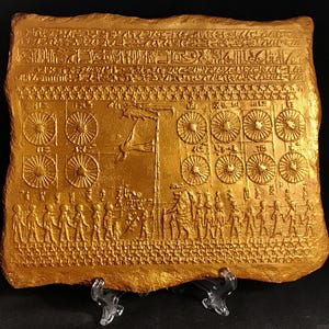 May include: A gold-coloured, rectangular relief sculpture depicting an ancient scene with figures, hieroglyphs, and circular motifs. The artwork is mounted on a clear stand, showcasing intricate details and historical themes.