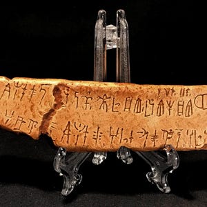 May include: A weathered, tan-colored artifact with etched markings, possibly ancient script. The rectangular piece is supported by a clear stand, suggesting display. The object has a broken edge, adding to its aged appearance.