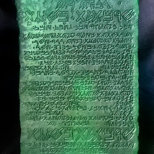 May include: A rectangular, green-tinted glass tablet with etched inscriptions. The text appears to be an ancient script, covering the entire surface. The tablet is supported by a clear, plastic stand, and the background is dark.