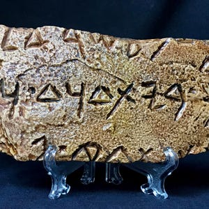 May include: A rectangular, weathered stone tablet with carved, stylized symbols. The tablet is a mix of tan and brown hues, with a rough, textured surface. It rests on a clear plastic display stand, suggesting it's a decorative or collectible item.