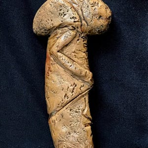 May include: A carved, tan-colored sculpture of a stylized figure. The sculpture has a rounded head and a textured, cylindrical body with carved details. The figure appears to be in a seated or reclining position. The sculpture is displayed against a dark background.