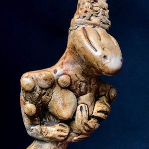 Op de afbeelding: Een bruinbeige keramische sculptuur van een gestileerde figuur met een afgeronde kop en lichaam. De figuur heeft een textuur met cirkelvormige en afgeronde uitsteeksels. Op een donkerblauwe achtergrond.