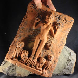Op de afbeelding: Een terracotta reliëfsculptuur met een gevleugelde vrouwelijke figuur die cirkelvormige objecten vasthoudt, geflankeerd door uilen en leeuwen. De figuur heeft een hoofdtooi en armbanden. De achtergrond heeft geëtste symbolen. De sculptuur wordt door een hand vastgehouden.