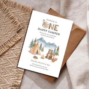 May include: A white invitation card with a camping theme. The text "Jackson is ONE HAPPY CAMPER" is displayed above an illustration of a tent, mountains, trees, a bear, and a fox. The invitation includes the date, time, and location.