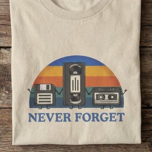 May include: Beige t-shirt with a retro design featuring a floppy disk, VHS tape, and cassette tape under a colorful sunset. The text "NEVER FORGET" is printed in blue below the graphics. The shirt is folded on a wooden surface.