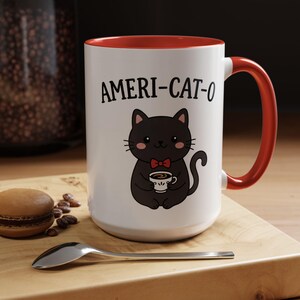 Puede incluir: Taza de cerámica blanca con interior y asa rojos. La taza presenta el texto "AMERI-CAT-O" sobre un gato negro de dibujos animados con pajarita roja y sosteniendo una taza de café. La taza está sobre una superficie de madera.