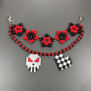 May include: A beaded chain featuring a top row of five star-shaped motifs made from red and black pony beads, connected to a lower strand of alternating red and black beads. Two charms hang from the lower strand: a white skull and a checkered heart.