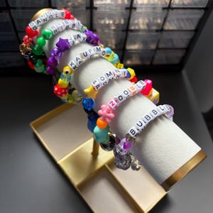 Könnte beinhalten: Eine Sammlung farbenfroher Perlenarmbänder mit weißen Buchstabenperlen, die Wörter wie "GANGLE", "JAX", "ZOOBLE" und "BUBBLE" bilden. Die Armbänder weisen verschiedene Formen und Farben auf, darunter Sterne, Herzen und Blumen.