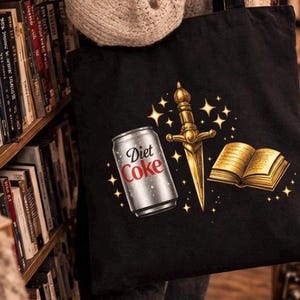 May include: Black tote bag featuring a Diet Coke can, a golden dagger, and a golden book with stars. The bag is held in front of a bookshelf filled with books. A candle and more books are on a table.