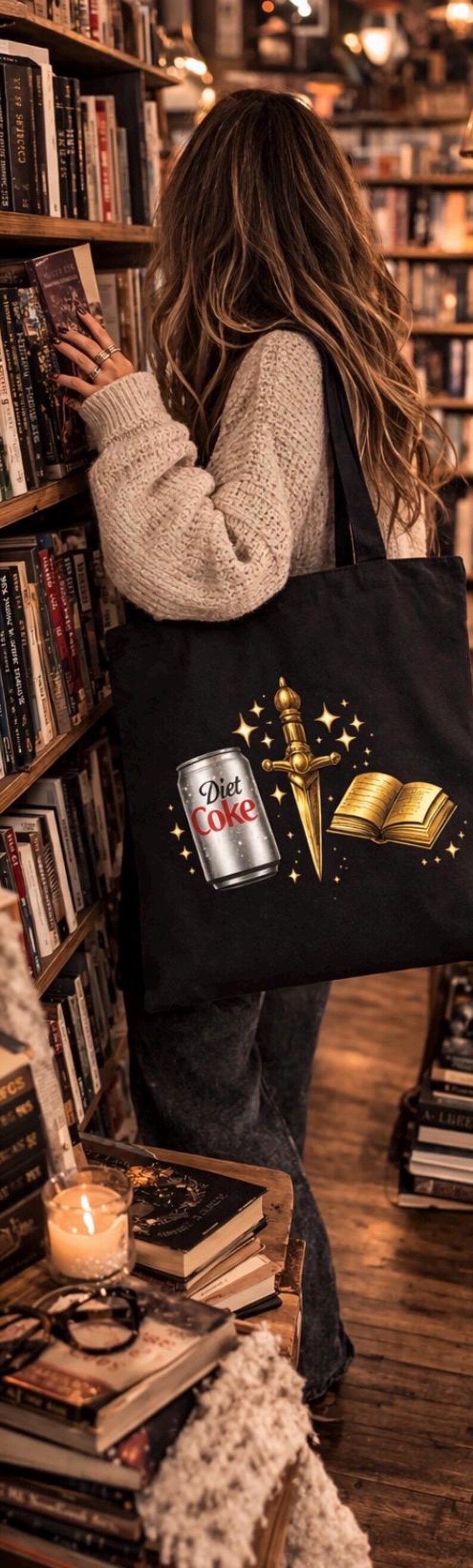 May include: Black tote bag featuring a Diet Coke can, a golden dagger, and a golden book with stars. The bag is held in front of a bookshelf filled with books. A candle and more books are on a table.