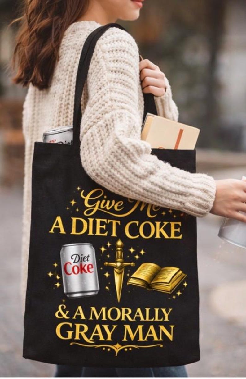 May include: Black tote bag with the text "Give Me A Diet Coke & A Morally Gray Man." The bag features a Diet Coke can, a sword, and an open book graphic. The bag is carried by a person wearing a cream-colored sweater.