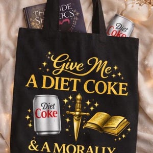 May include: Black tote bag with the text "Give Me A DIET COKE & A MORALLY GRAY MAN" in gold lettering. The bag features illustrations of a Diet Coke can, a dagger, and an open book. Two books are visible inside the bag.