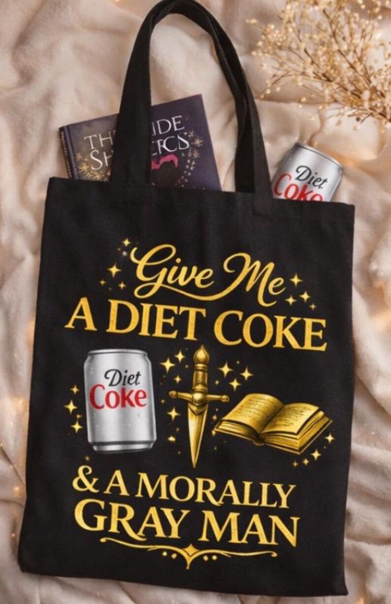May include: Black tote bag with the text "Give Me A DIET COKE & A MORALLY GRAY MAN" in gold lettering. The bag features illustrations of a Diet Coke can, a dagger, and an open book. Two books are visible inside the bag.