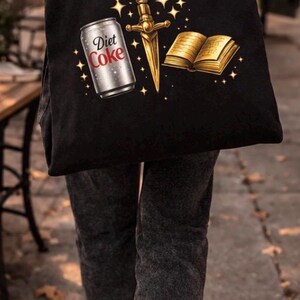 May include: Black tote bag featuring a Diet Coke can, a golden dagger, and an open book with gold-colored pages. The bag is carried by a person walking on a sidewalk.