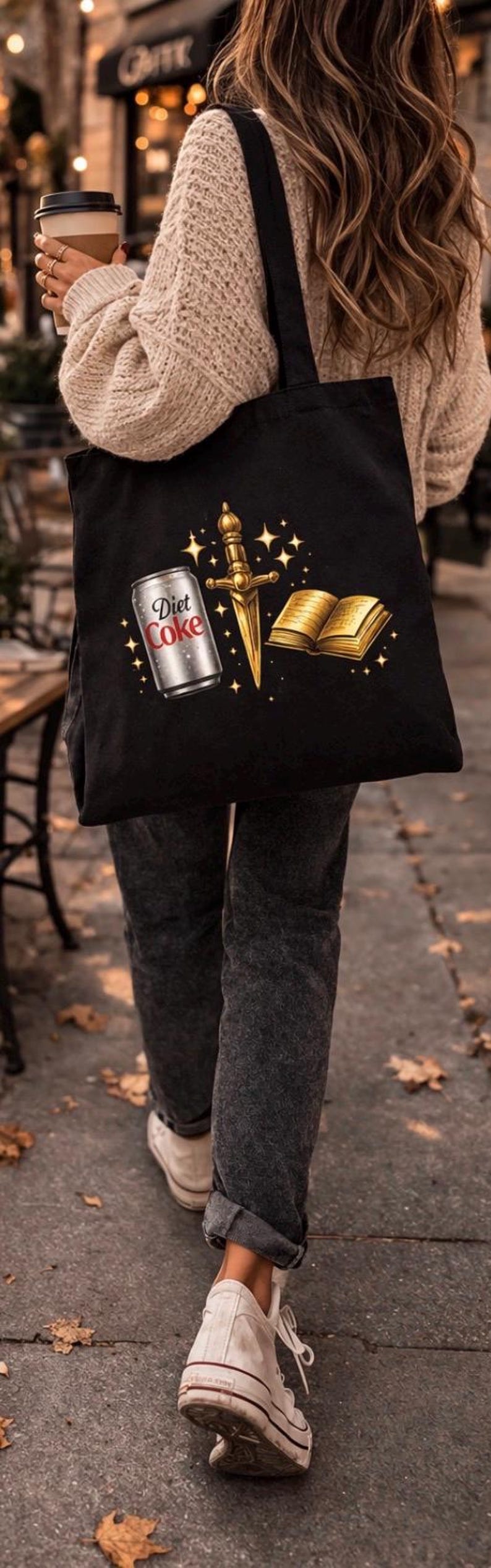 May include: Black tote bag featuring a Diet Coke can, a golden dagger, and an open book with gold-colored pages. The bag is carried by a person walking on a sidewalk.