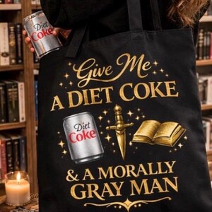 May include: Black tote bag with the text "Give Me A Diet Coke & A Morally Gray Man" in gold lettering. The design includes a Diet Coke can, a dagger, and a book. A Diet Coke can is held in the background.