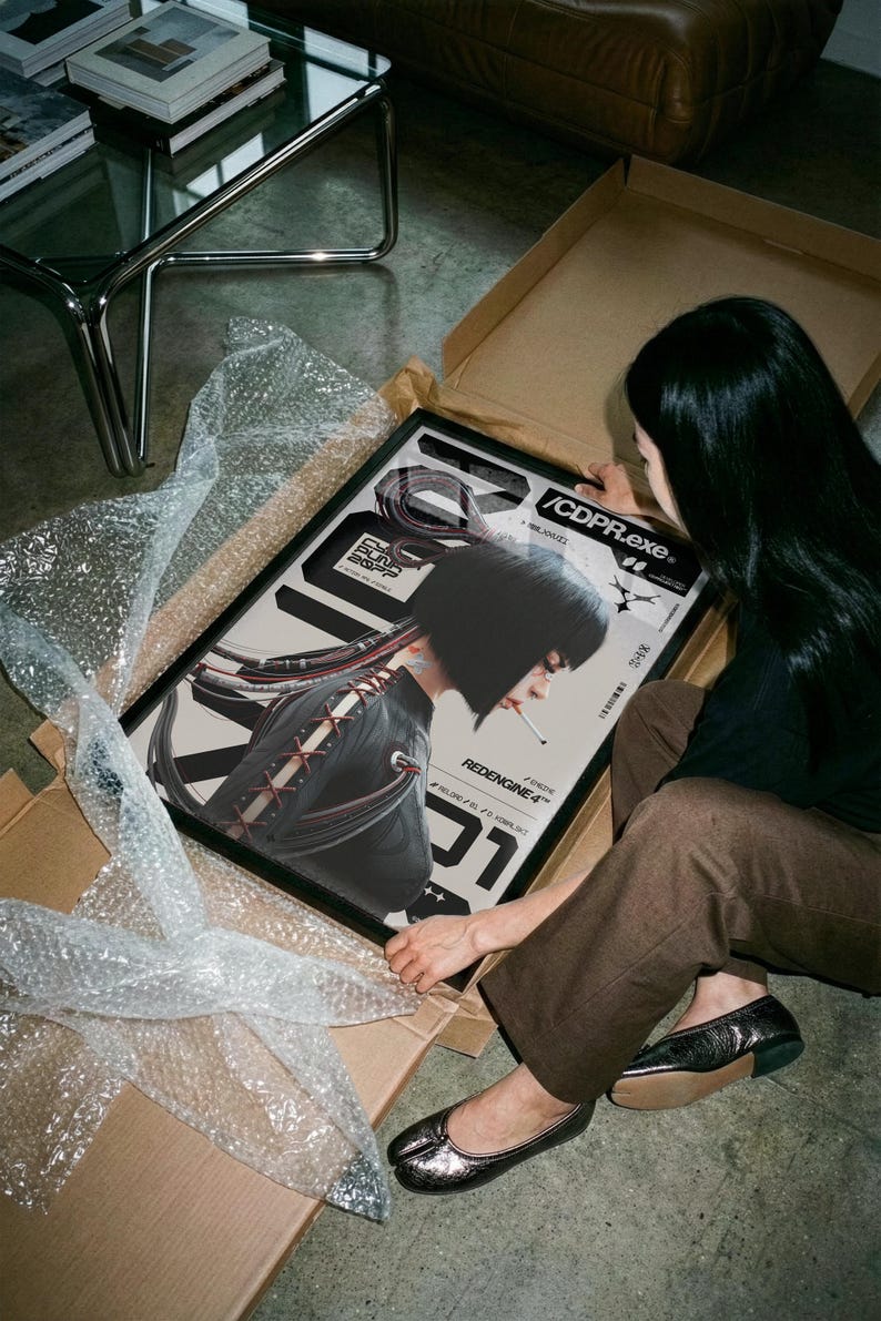 Op de afbeelding: Een ingelijste poster met een futuristische illustratie van een persoon met een kort zwart kapsel, die een sigaret rookt. De poster heeft een zwart frame en wordt uitgepakt uit een kartonnen doos. De tekst "/CDPR.exe" en "REDENGINE4" is zichtbaar.