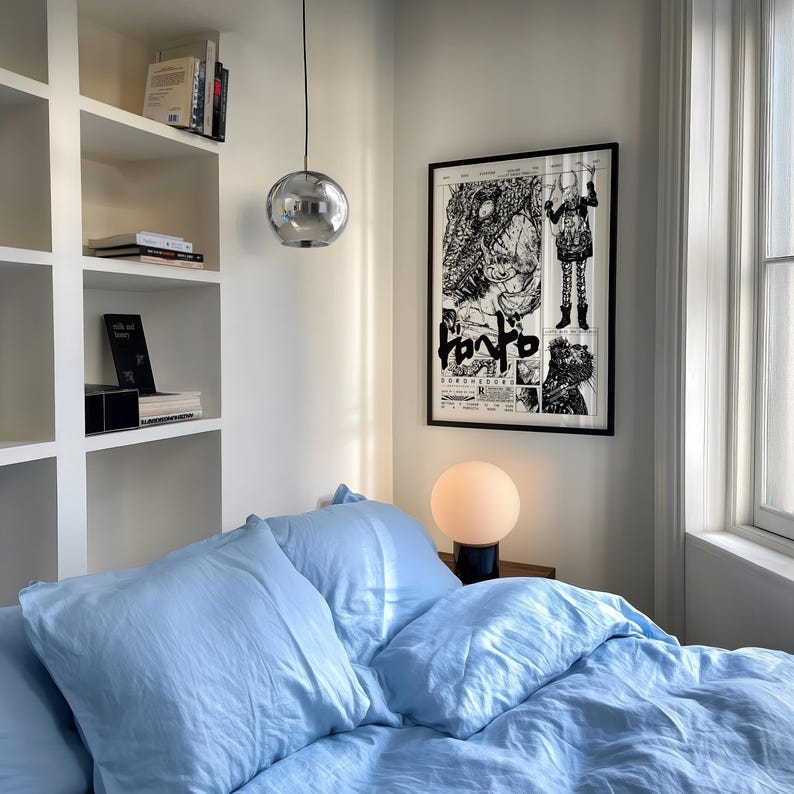 May include: A bedroom interior with a light blue bed set. A black framed poster with black and white illustrations hangs on the wall. A spherical chrome pendant light hangs from the ceiling. A small, round, white lamp sits on a wooden nightstand.
